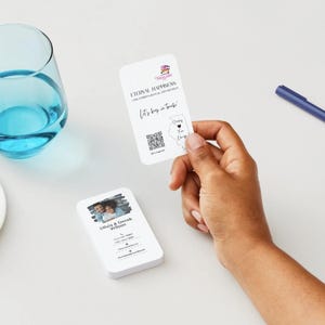 Peut inclure: Cartes de visite blanches aux coins arrondis. L'une est tenue &agrave; la main, affichant du texte et un code QR. Une autre carte montre une photo de couple et des coordonn&eacute;es. Un verre de liquide bleu et des macarons sont &eacute;galement visibles.