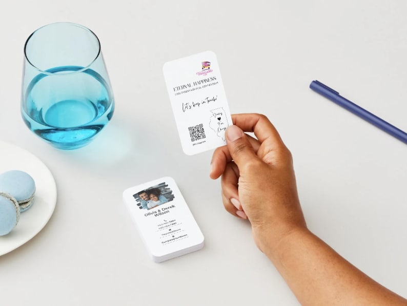 Peut inclure: Cartes de visite blanches aux coins arrondis. L'une est tenue &agrave; la main, affichant du texte et un code QR. Une autre carte montre une photo de couple et des coordonn&eacute;es. Un verre de liquide bleu et des macarons sont &eacute;galement visibles.