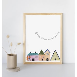 May include: A framed art print featuring a row of colorful, illustrated houses with a couple in front of the first house. The text "Queremos mucho a nuestros precarizos" is written above the houses in cursive script. The frame is light wood.