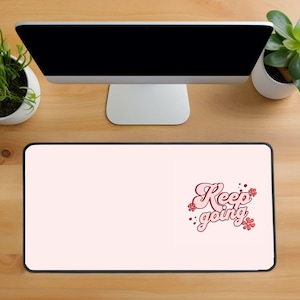 Puede incluir: Alfombrilla de ratón rectangular con fondo rosa claro y las palabras "Keep going" en una fuente retro con detalles florales. Un monitor de ordenador y plantas en macetas son visibles en el fondo.