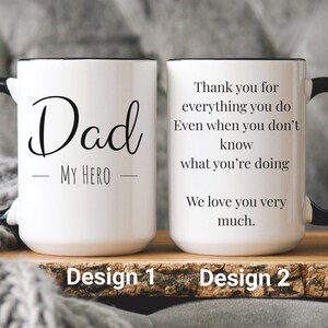 Op de afbeelding: Twee witte keramische mokken met zwarte handvatten en randen. Eén mok zegt "Dad MY HERO" in zwart schrift. De andere mok zegt "Thank you for everything you do... We love you very much." Design 1 en Design 2 zijn gelabeld.