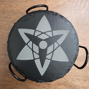 May include: A round, black slate coaster with a gray, stylized star design. The coaster is held in a black metal holder with two curved handles. The design is centered on the coaster.