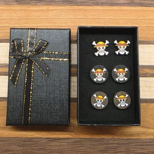 May include: A black gift box with a gold bow and a matching open box containing six skull and crossbones earrings. The earrings have a yellow hat and are on a black background.
