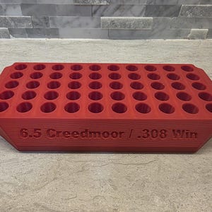 May include: A red rectangular ammunition loading block featuring fifty circular holes arranged in five rows of ten. The front side of the block is embossed with the text 6.5 Creedmoor / .308 Win, indicating the compatible cartridge types.
