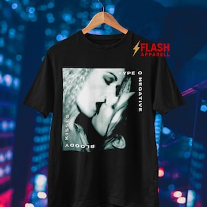 Może przedstawiać: Czarna koszulka z szarą grafiką przedstawiającą dwie całujące się osoby. Słowa "BLOODY KISSES" są wydrukowane pionowo po lewej stronie, a "TYPE O NEGATIVE" po prawej. Koszulka ma również napis "FLASH APPAREL" w prawym górnym rogu.