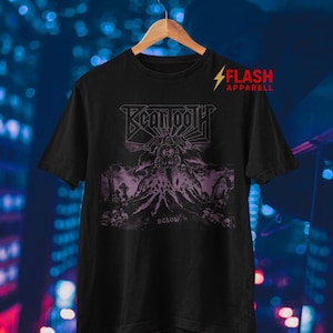 May include: Black t-shirt with a purple graphic featuring the band name "Behemoth" and a detailed illustration of a figure with a dark, gothic theme. The text "BELOW" is also visible. The shirt is hanging on a wooden hanger.