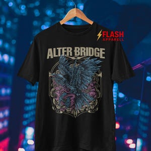 May include: Black t-shirt featuring the band name "ALTER BRIDGE" above a detailed graphic of a bird with outstretched wings, surrounded by floral and serpentine elements. The shirt includes the "FLASH APPAREL" logo.