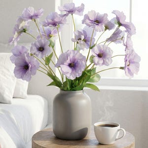 May include: A display of lavender-coloured flowers in a grey vase on a wooden table. A white coffee cup is beside the vase. The flowers have dark centres and green stems. The scene is illuminated by natural light.