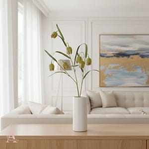May include: A white cylindrical vase holds a decorative plant with green and brown bell-shaped flowers and long green leaves. The vase sits on a light wood table in front of a beige sofa and a painting with gold accents.