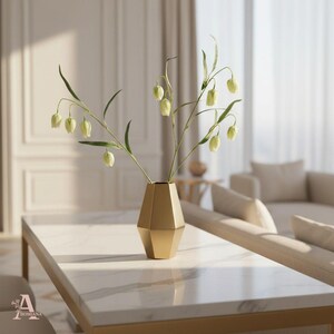 May include: A gold geometric vase holding green and white flowers sits on a white marble coffee table. The vase has a unique, angular design. The table has a gold frame. The background includes a beige sofa and a window with sheer curtains.
