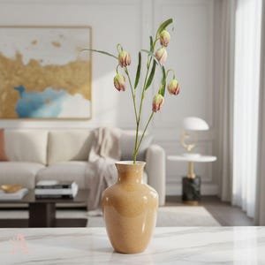 May include: A tan ceramic vase holds a floral arrangement with green stems and bell-shaped flowers in shades of pink and green. The vase sits on a white marble surface, with a living room setting in the background.