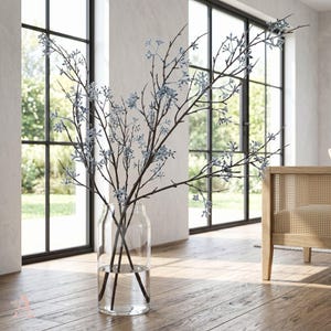 Può includere: Un vaso di vetro trasparente contiene diversi rami marrone scuro con delicati accenti floreali blu chiaro. Il vaso è posto su un pavimento in legno, con una finestra e una sedia di legno sullo sfondo. L'arrangiamento aggiunge un tocco naturale.