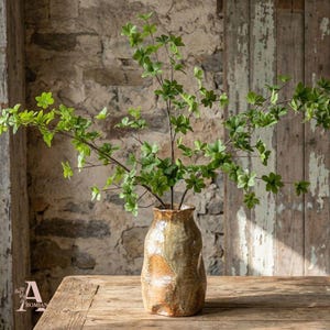 Könnte beinhalten: Eine strukturierte Keramikvase in Braun- und Beigetönen, gefüllt mit grünen, belaubten Zweigen. Die Vase steht auf einem Holztisch, vor einem Hintergrund aus Stein und verwittertem Holz.