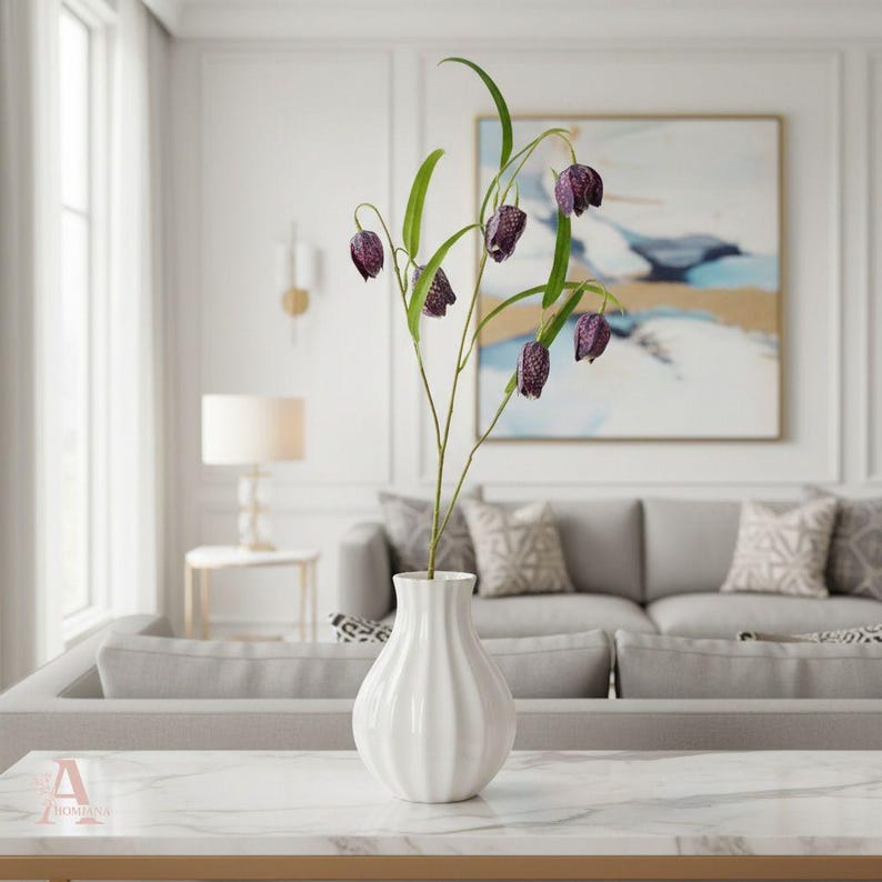 May include: A white, ribbed ceramic vase holding a stem of purple and green flowers. The flowers have a bell-like shape with a speckled pattern. The vase sits on a marble-topped table in a living room setting.