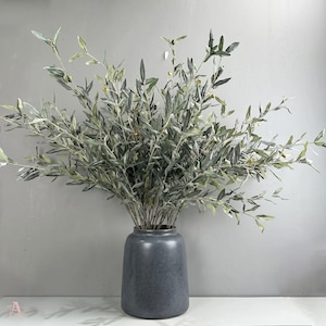 Puede incluir: Un arreglo decorativo de ramas de olivo artificiales con hojas verdes y grises y pequeños acentos en forma de aceituna. Las ramas están dispuestas en un jarrón de cerámica gris, creando una estética natural y elegante.