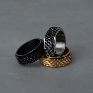 May include: Three textured metal rings in black, silver, and gold. The rings have a scale-like pattern and are stacked on top of each other. The rings are on a gray surface.
