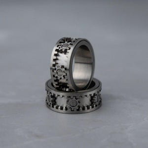 May include: Two silver-colored rings with a gear design. The rings are stacked on top of each other, with the top ring in focus. The gear design is etched around the outer band of the rings, creating a mechanical aesthetic. The rings appear to be made of metal.