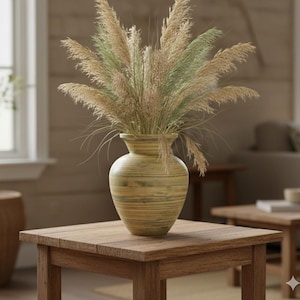 May include: A decorative vase with a wide base and narrow neck, filled with dried ornamental grasses. The vase is a light green and yellow color, and sits on a square wooden table. The room has a neutral color palette.
