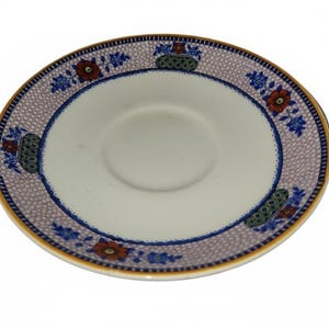May include: A white ceramic saucer with a central indentation. The rim features a repeating pattern of blue floral and geometric designs on a lavender background. A gold-colored band edges the saucer.