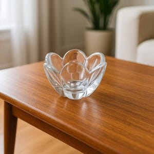 May include: A clear glass bowl with a flower-like design, resting on a wooden table. The bowl has a central well and six petal-shaped sections. The table is a warm, medium brown color, and the background is blurred.