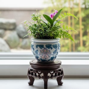 May include: A potted plant with green foliage and a purple orchid flower sits in a decorative ceramic pot with blue and pink floral patterns. The pot rests on a dark brown, carved wooden stand with three legs, placed near a window.