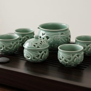 May include: A celadon green tea set with intricate floral carvings. The set includes a teapot, a lidded container, and four small cups, all arranged on a dark wooden tray with smooth, black stones. A serene and elegant tea ceremony set.