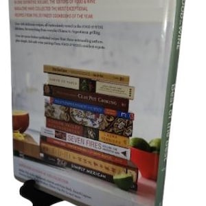 May include: A stack of cookbooks, including "Seven Fires" and "Simply Mexican", is displayed on a book stand. The cover of the book on the front says "Food & Wine Best of the Best". The books are on a white surface.