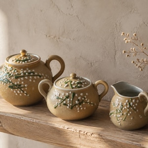 May include: A ceramic tea set in a textured mustard yellow glaze featuring a raised white and green floral pattern. The set includes a lidded teapot, a lidded sugar bowl with two handles, and a creamer pitcher, all decorated with matching sprigs.