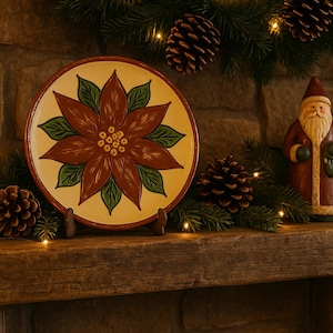 May include: A festive holiday display featuring a hand-painted plate with a poinsettia design, a Santa Claus figurine, pine cones, and a lit candle. The plate is supported by a wooden stand, and the scene is decorated with evergreen branches and fairy lights.