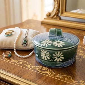 May include: A round, lidded ceramic trinket box in shades of deep green and blue with white daisy patterns, accompanied by a white velvet pillow holding a pearl necklace, a silver pendant, and several rings, plus a separate silver flower brooch.