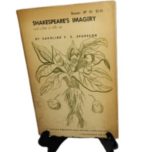 May include: A vintage book titled "Shakespeare's Imagery" by Caroline F. E. Spurgeon. The cover is beige with a botanical illustration and text. The book is propped up on a black display stand.