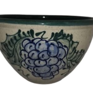 May include: A ceramic bowl with a cream-colored exterior and a dark green rim. The bowl is decorated with a blue and green grape design. The bowl is ideal for serving food or as a decorative piece.