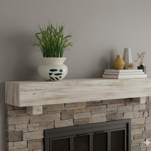 May include: A white vase with green leaf-shaped designs holds tall green grass, placed on a distressed white wooden mantel. The mantel is above a stone fireplace, with books and decorative objects.