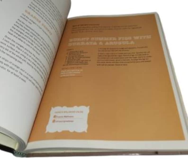 May include: Open cookbook pages with text and a recipe for Burnt Summer Figs with Burrata & Arugula. The right page is tan with the recipe title in brown. The left page has text about oranges. A white label with social media handles is on the right page.