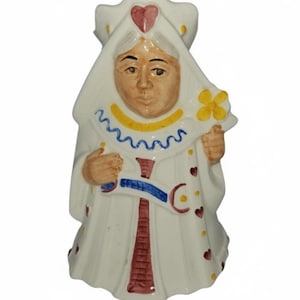 May include: A ceramic figurine of the Queen of Hearts from Alice in Wonderland. The queen is dressed in a white gown with red, blue, and yellow accents. A red heart is on her crown, and she holds a yellow flower.