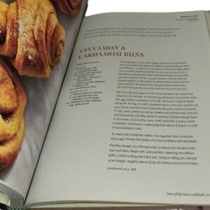 May include: An open cookbook displays a recipe for cinnamon and cardamom buns. The left page shows several golden-brown, baked buns. The right page features the recipe title and text. The book is on a white surface.