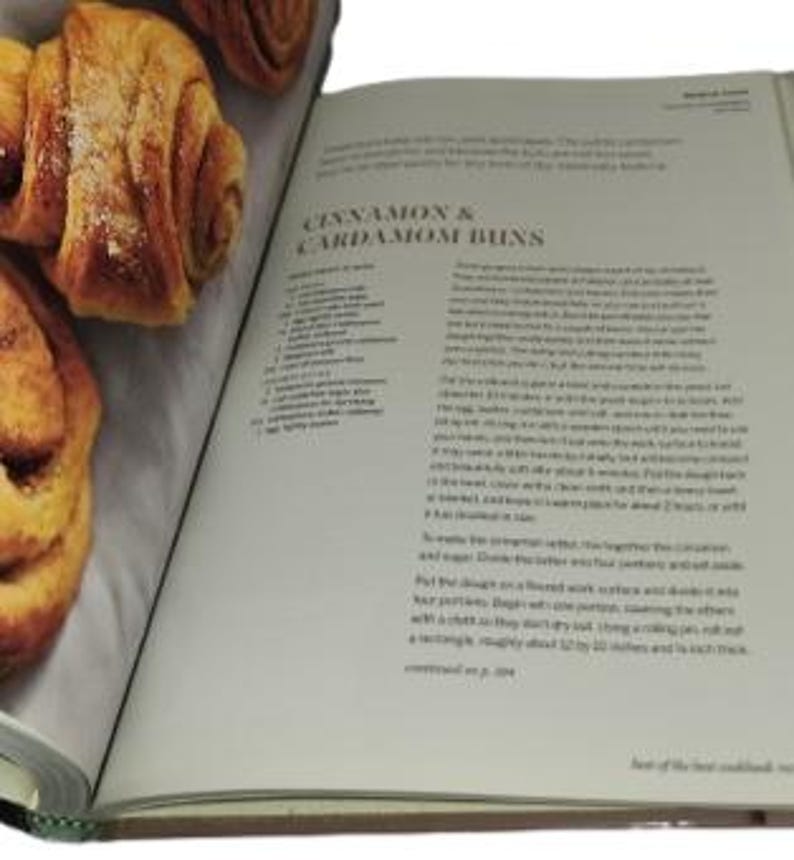 May include: An open cookbook displays a recipe for cinnamon and cardamom buns. The left page shows several golden-brown, baked buns. The right page features the recipe title and text. The book is on a white surface.