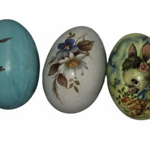May include: Three decorative Easter eggs. One is light blue with a rabbit illustration, another is white with floral designs, and the third is green with a donkey illustration. The eggs are oval-shaped.