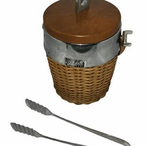 May include: Vintage ice bucket with a brown lid and woven basket base, accented with a silver band and handle. Includes silver ice tongs. The ice bucket has the text "Likewood" on the front.