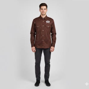 May include: A brown button-down shirt with two chest pockets and a small white emblem. The shirt has long sleeves and is paired with dark gray pants and black shoes. The person is standing in front of a plain white background.