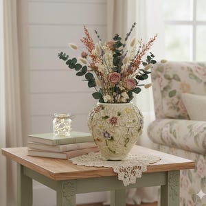 May include: A cream-colored vase with floral relief, filled with dried flowers, sits on a wooden table. The table also holds books, a jar of fairy lights, and a lace doily. A floral-patterned armchair is in the background.