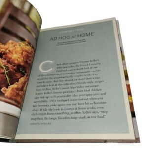 May include: Open cookbook with the title "AD HOC AT HOME" on a light blue page. The text describes family-style recipes from Ad Hoc, Keller's casual Napa Valley restaurant. The left page shows a close-up of fried food.