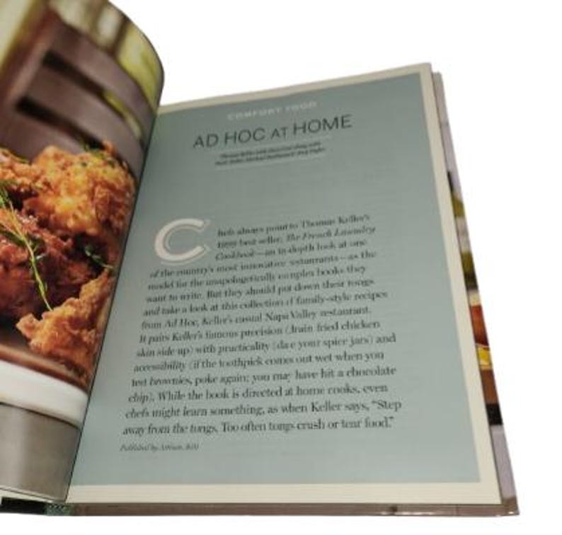 May include: Open cookbook with the title "AD HOC AT HOME" on a light blue page. The text describes family-style recipes from Ad Hoc, Keller's casual Napa Valley restaurant. The left page shows a close-up of fried food.
