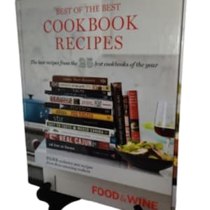 May include: A cookbook titled "Best of the Best Cookbook Recipes" with a stack of cookbooks visible. The cover text is in red. The book is propped up on a black stand. The words "Food & Wine" are visible on the bottom.