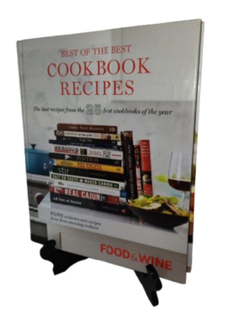 May include: A cookbook titled "Best of the Best Cookbook Recipes" with a stack of cookbooks visible. The cover text is in red. The book is propped up on a black stand. The words "Food & Wine" are visible on the bottom.