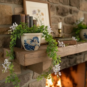 May include: A rustic mantelpiece with a wooden shelf displays a cream-colored ceramic pot with blue floral designs, filled with greenery and white flowers. A framed botanical print and antique books are also on the shelf, creating a cozy, vintage aesthetic.