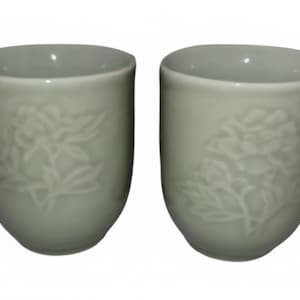 May include: Two pale green ceramic mugs with a floral design. Each mug has a handle and a detailed embossed peony flower pattern. The mugs are a soft, muted green color.
