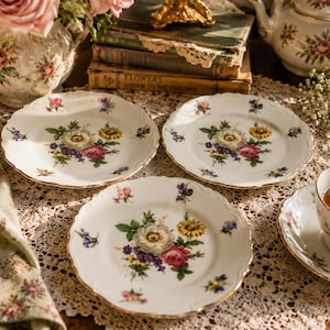 May include: Three white porcelain plates with scalloped edges and gold trim, each featuring a central floral bouquet of pink, yellow, and purple flowers with green leaves, arranged on a white lace tablecloth with matching floral tea service.