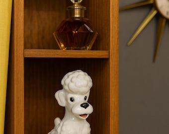 May include: A wooden shelf unit displays a vintage white ceramic poodle figurine and a decorative perfume bottle. The bottle has a faceted glass body and a crystal stopper. A gold starburst clock is visible in the background.