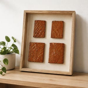 May include: A rectangular wooden shadow box frame containing four rectangular, textured, burnt-orange ceramic tiles arranged in a two-by-two grid against a light beige fabric backing. The tiles feature a glossy, organic, crackled surface pattern.